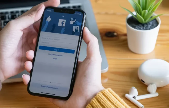 A woman in a yellow jumper holding a smartphone that's showing the Facebook log in page