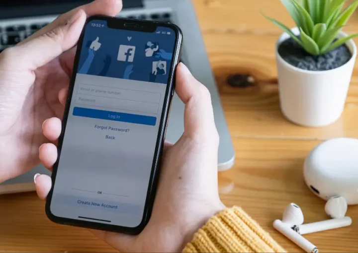 A woman in a yellow jumper holding a smartphone that's showing the Facebook log in page